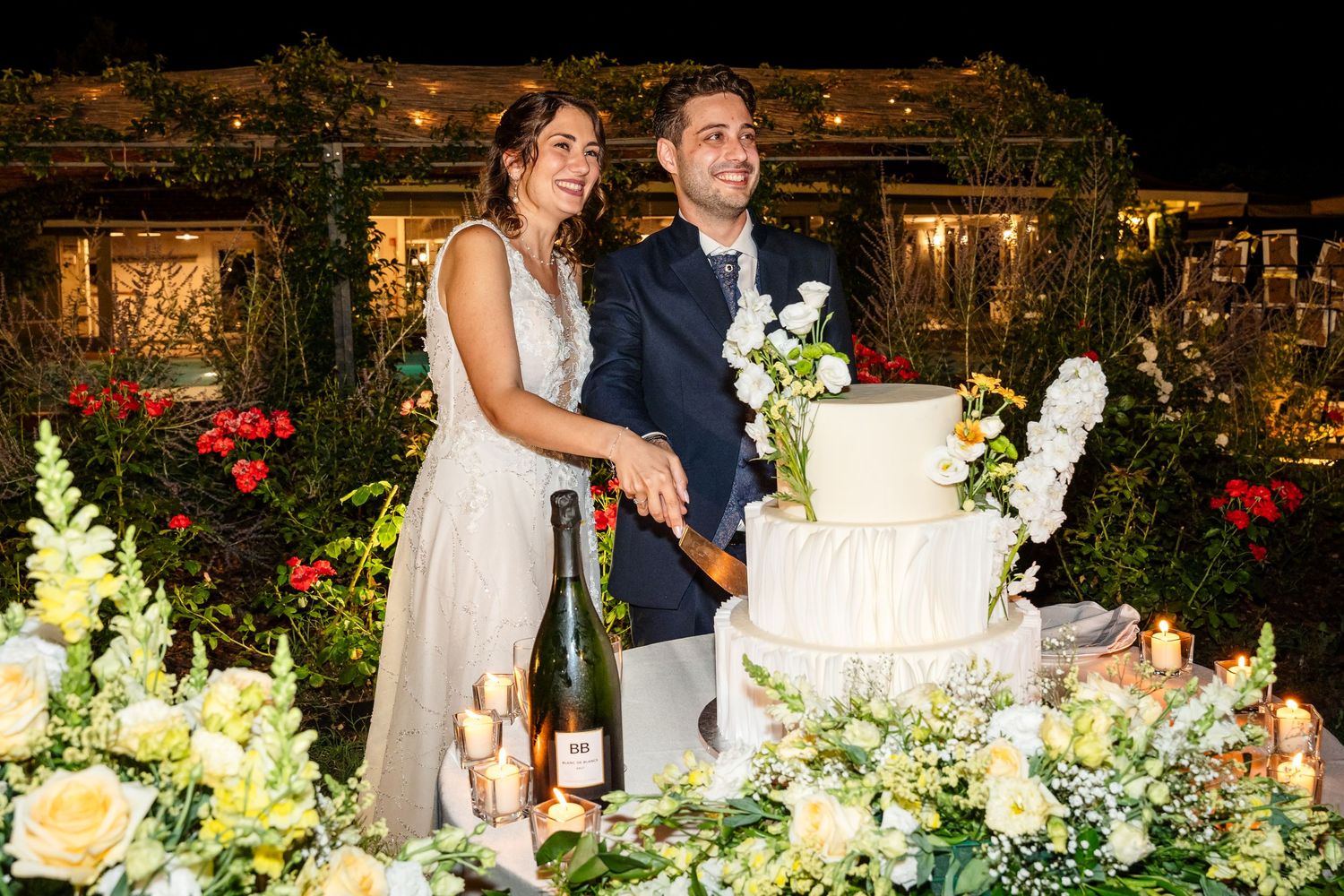 Bride and groom smiling while cutting a white floral wedding cake outdoors at night with champagne bottle nearby.