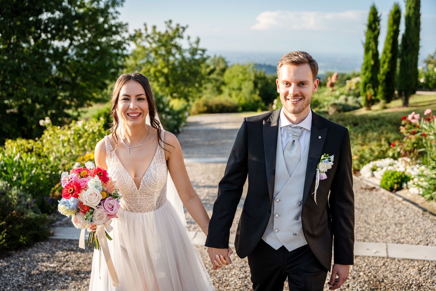 Happy newlyweds walk hand in hand outdoors, bride holding colorful bouquet in elegant white gown, groom in navy suit.