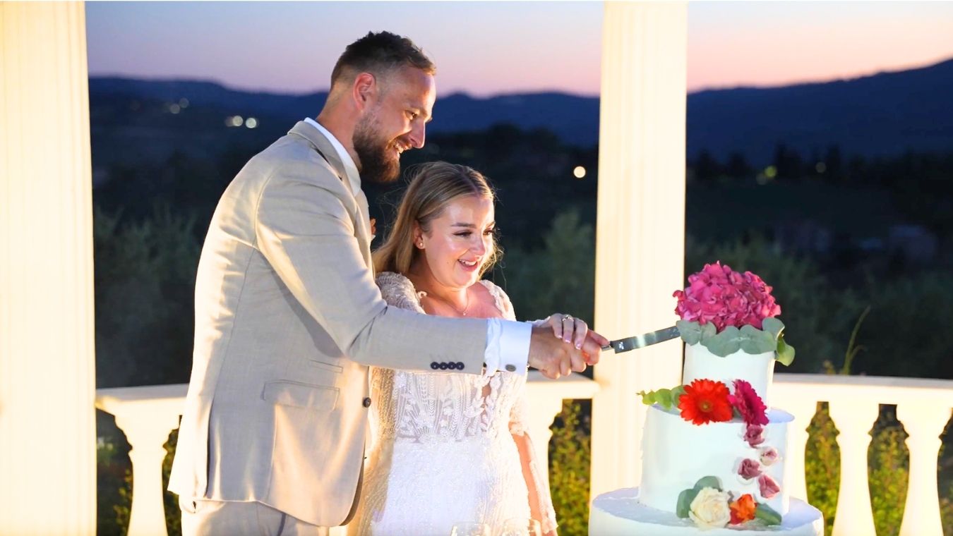 Couple cutting wedding cake at sunset on elegant white columned venue with colorful floral decorations and mountain views.