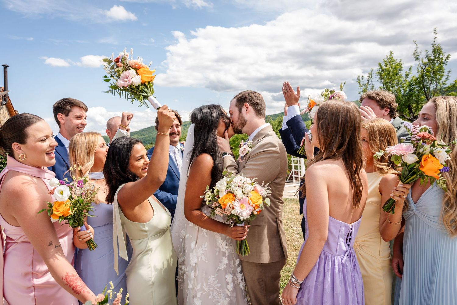 Wedding party celebrating outdoors, bride and groom kissing surrounded by bridesmaids holding colorful bouquets under blue sky.