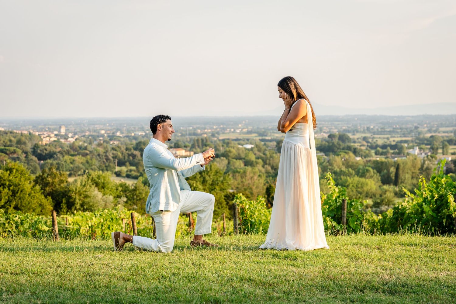 Man proposing on one knee to woman in vineyard at sunset with scenic landscape in background.