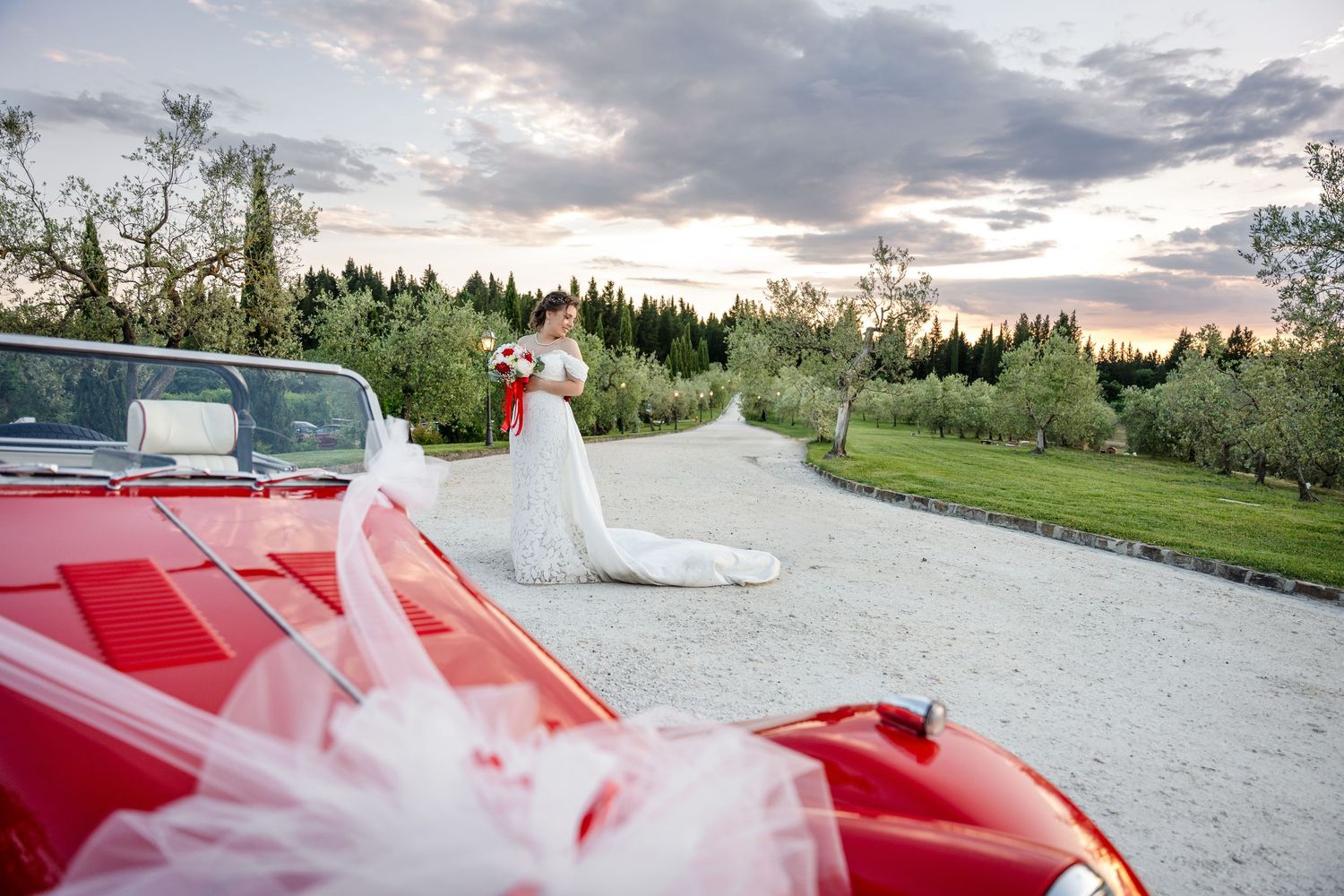 Bride in white gown stands near a red convertible car on a gravel path surrounded by lush greenery at sunset.