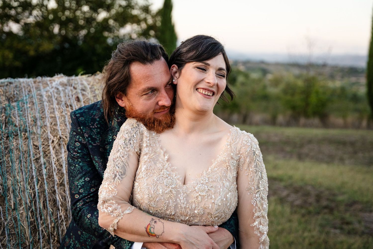 A smiling bride in lace dress embraced by groom with red beard near hay bale in rustic outdoor wedding setting.