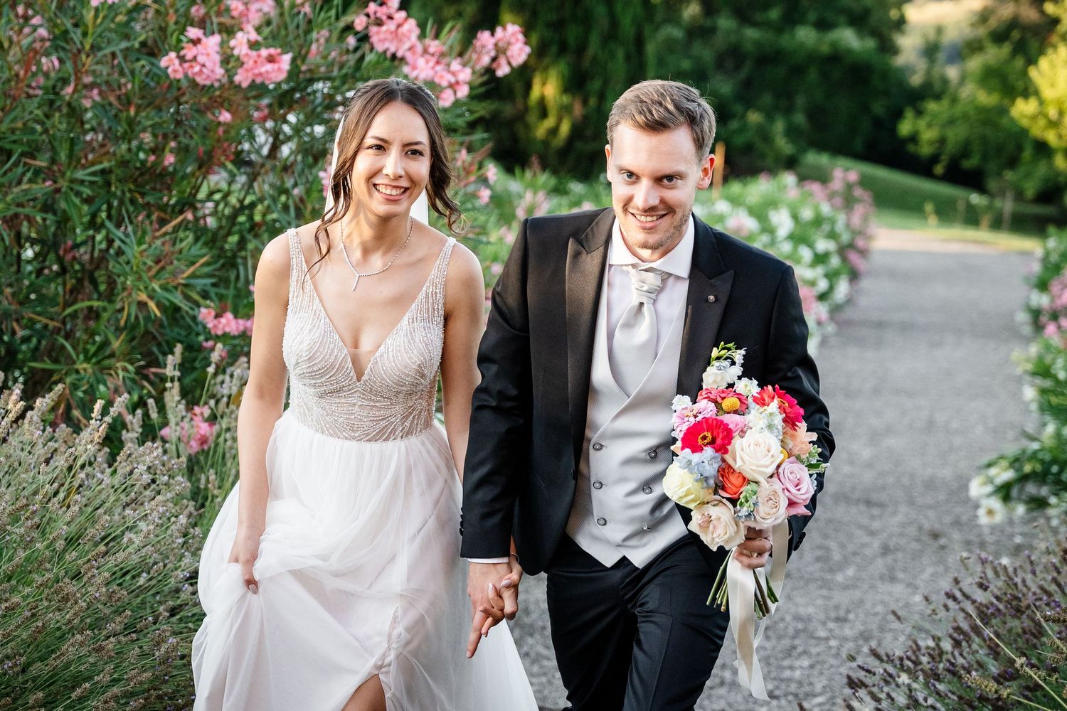 Smiling bride and groom walking hand in hand through a lush garden, groom holding a colorful bouquet.