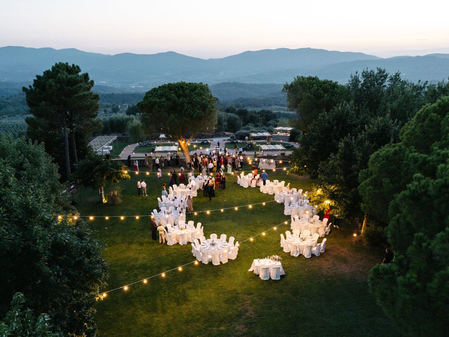Aerial view of an outdoor evening wedding reception with string lights arranged in a circular pattern on a hilltop lawn.
