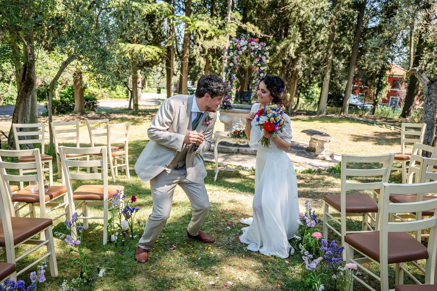 A scenic outdoor wedding venue with white chairs arranged in rows beneath shady trees and white decorative elements.