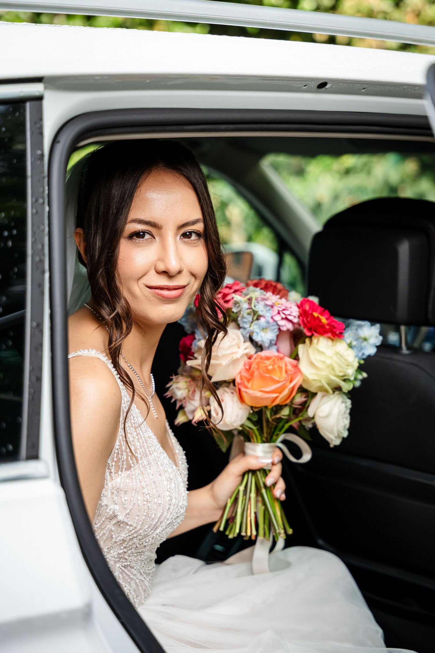 Bride in white gown holding colorful bouquet sitting in car window on wedding day.