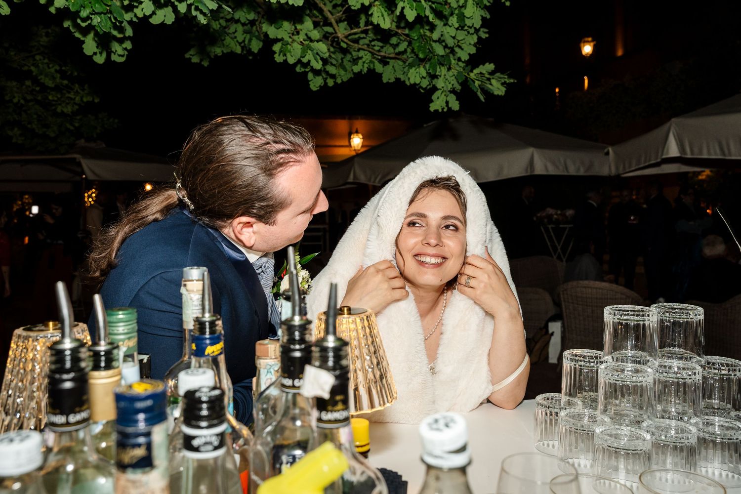 Smiling bride wrapped in veil sits with groom at outdoor evening wedding reception bar table.