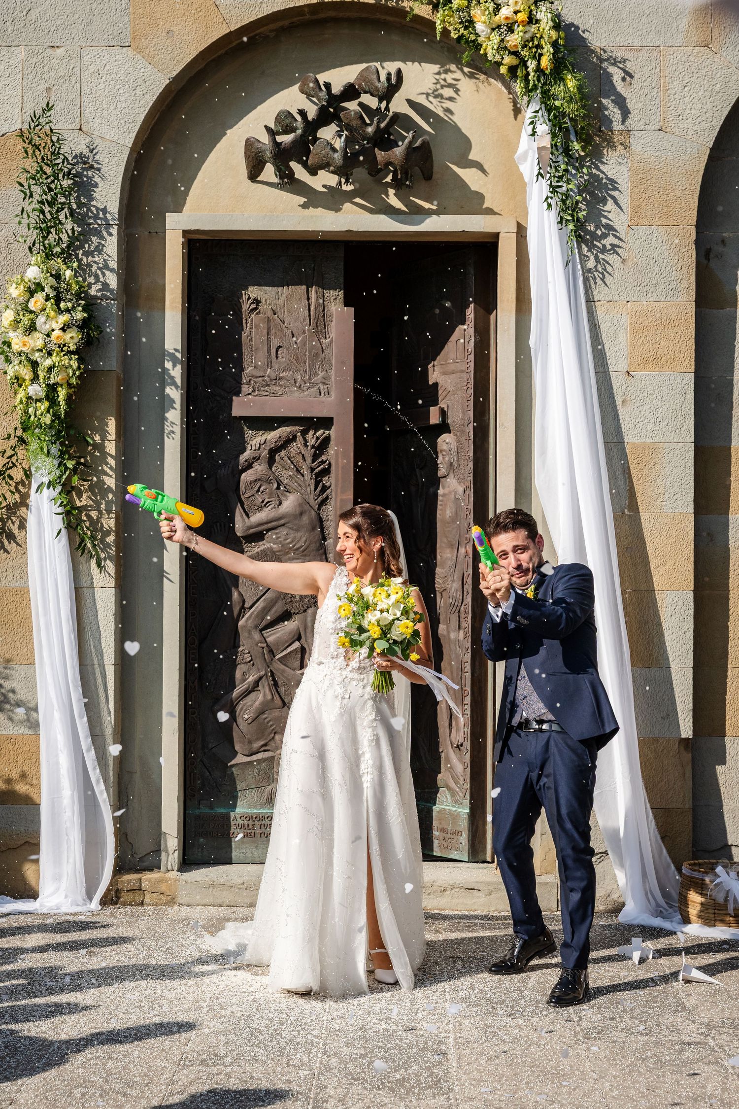Joyful newlyweds celebrate outside church doors, bride in white gown holding bouquet and groom in navy suit, both wielding water guns.