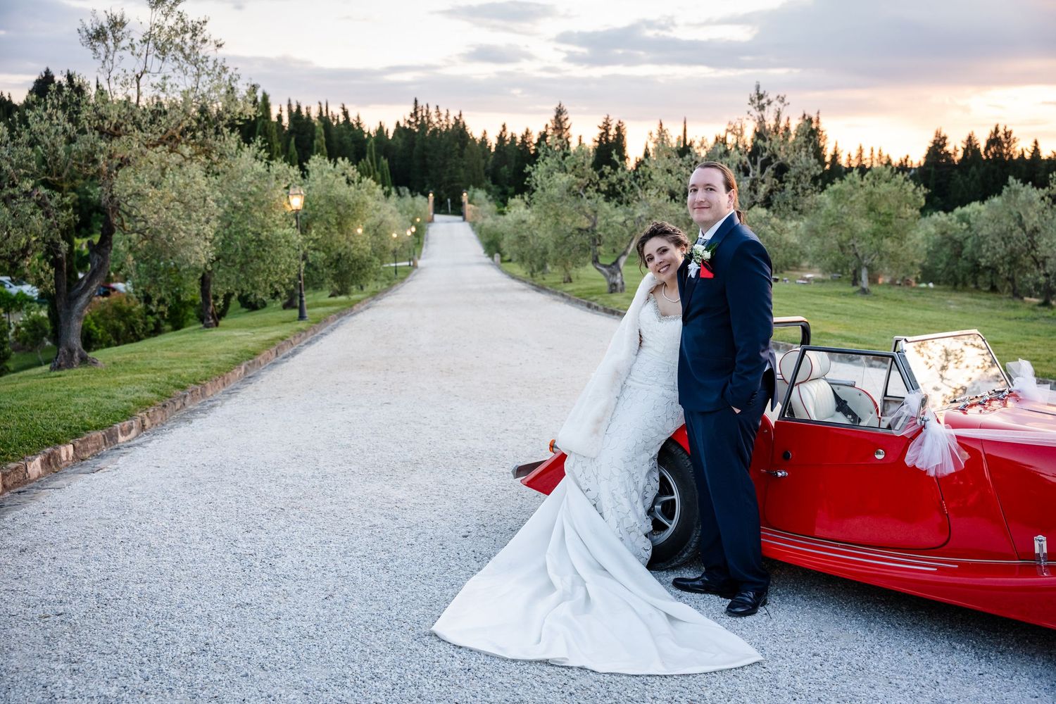 Elegant bride and groom posing beside a classic red convertible on a tree-lined gravel driveway at sunset.