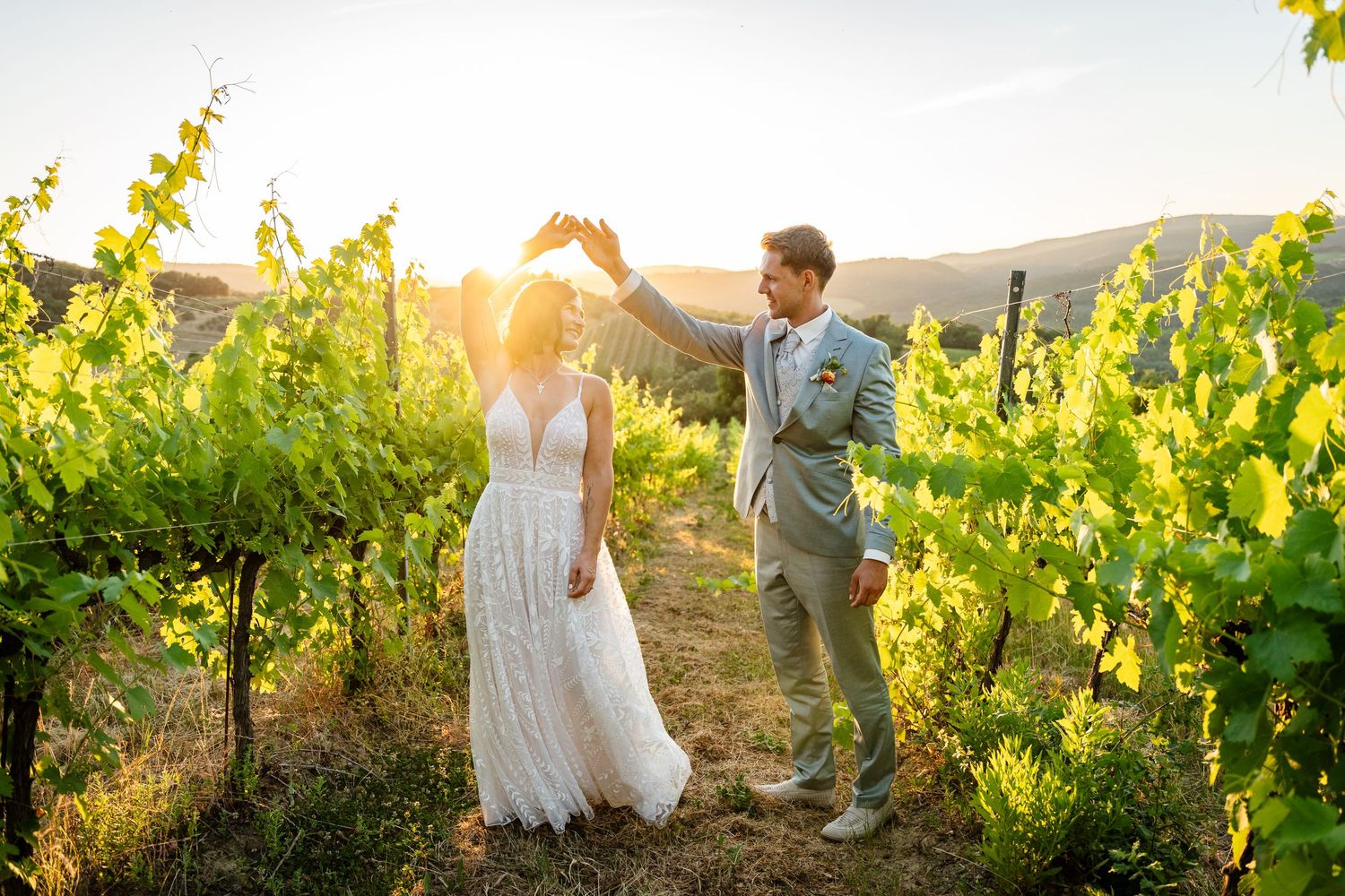 Bride and groom dancing at golden hour in a lush vineyard, sun setting behind rolling hills.