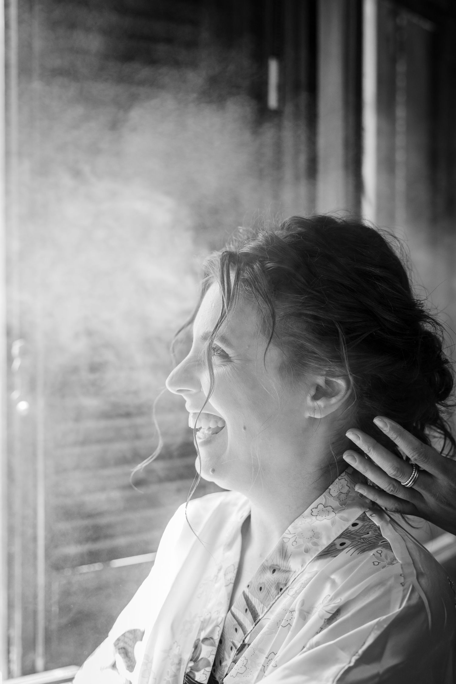 Black and white portrait of a smiling bride getting ready, with soft light streaming through a window.