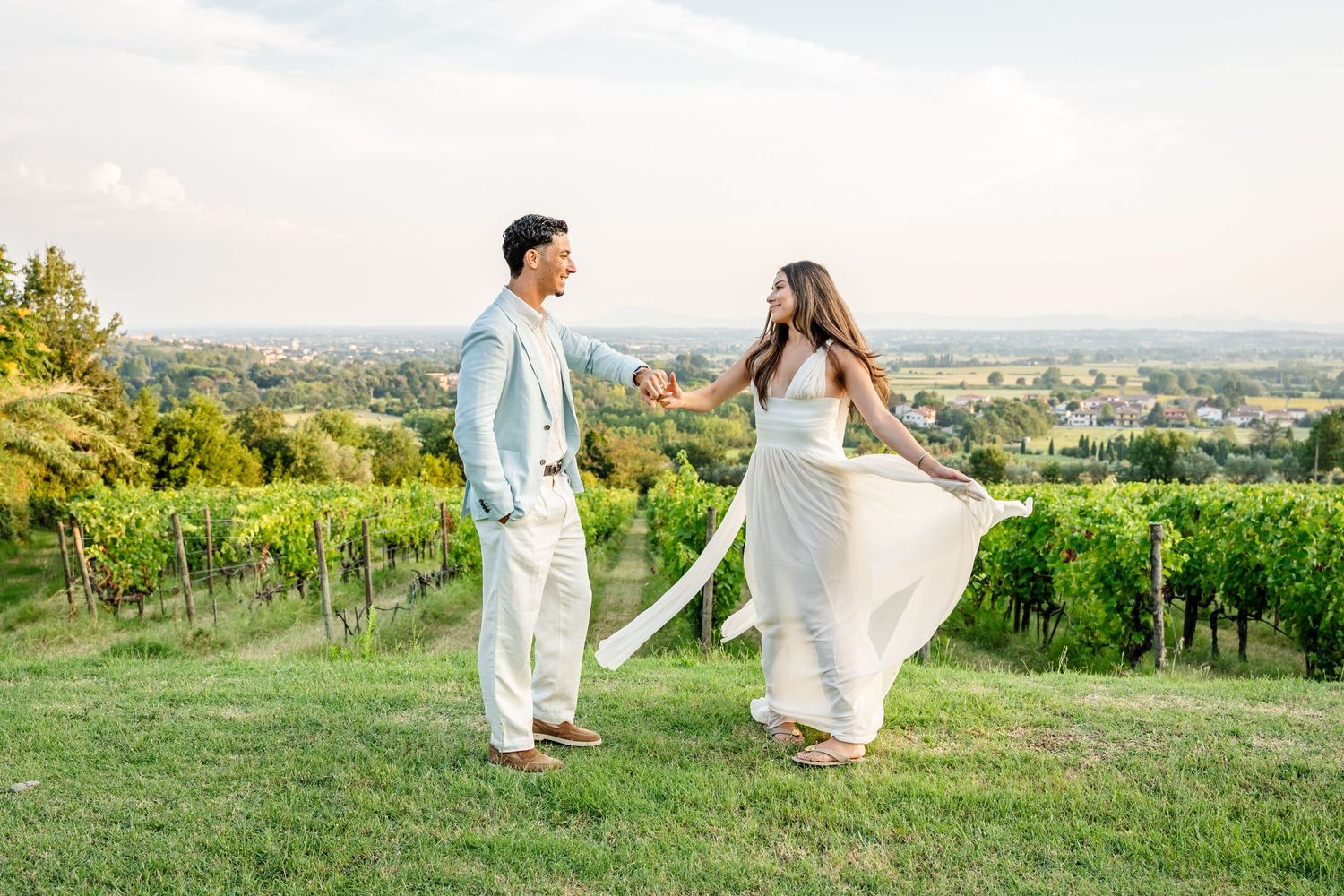 Couple dancing in vineyard, woman in flowing white dress, man in light blue suit, green hills backdrop.