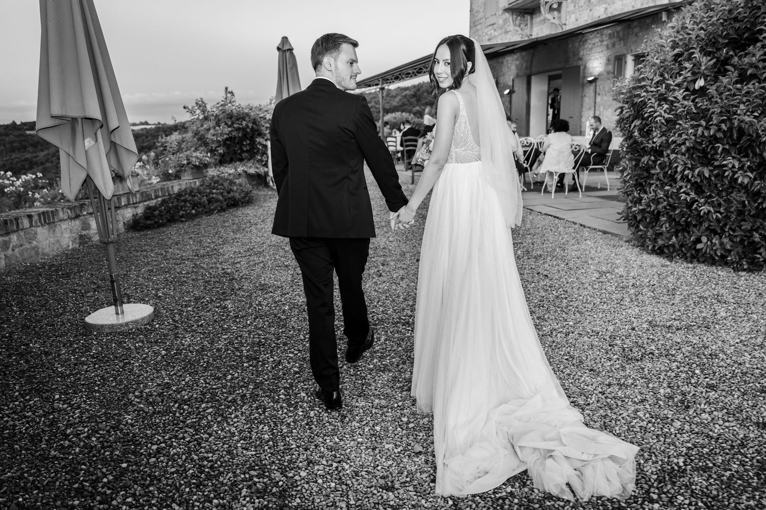 Black and white photo of bride and groom walking hand in hand through outdoor wedding venue grounds.