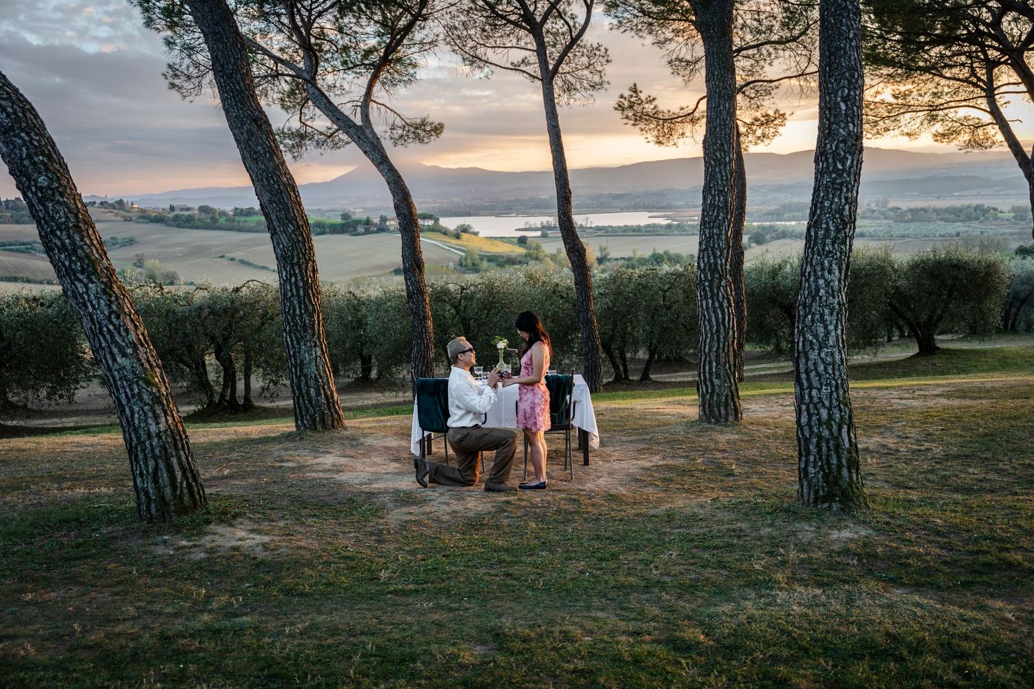 Group enjoys a scenic sunset picnic among tall pine trees on a grassy hillside overlooking a misty valley.