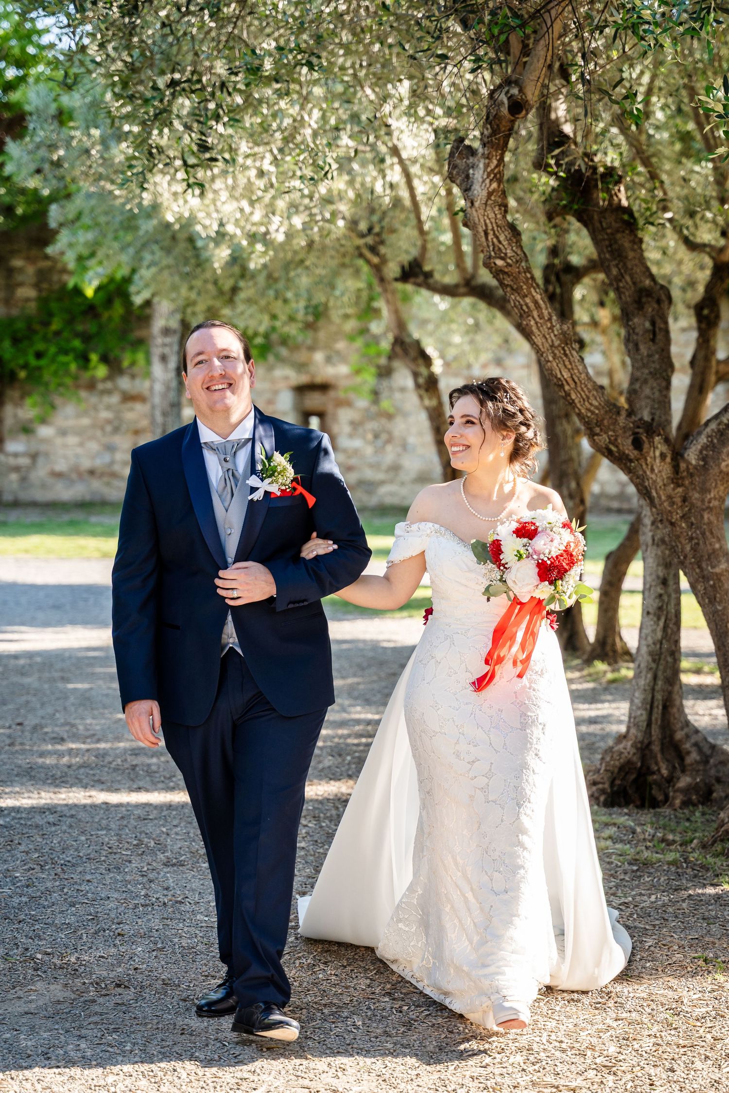 Smiling bride and groom walk together under olive trees at a Tuscan wedding venue.