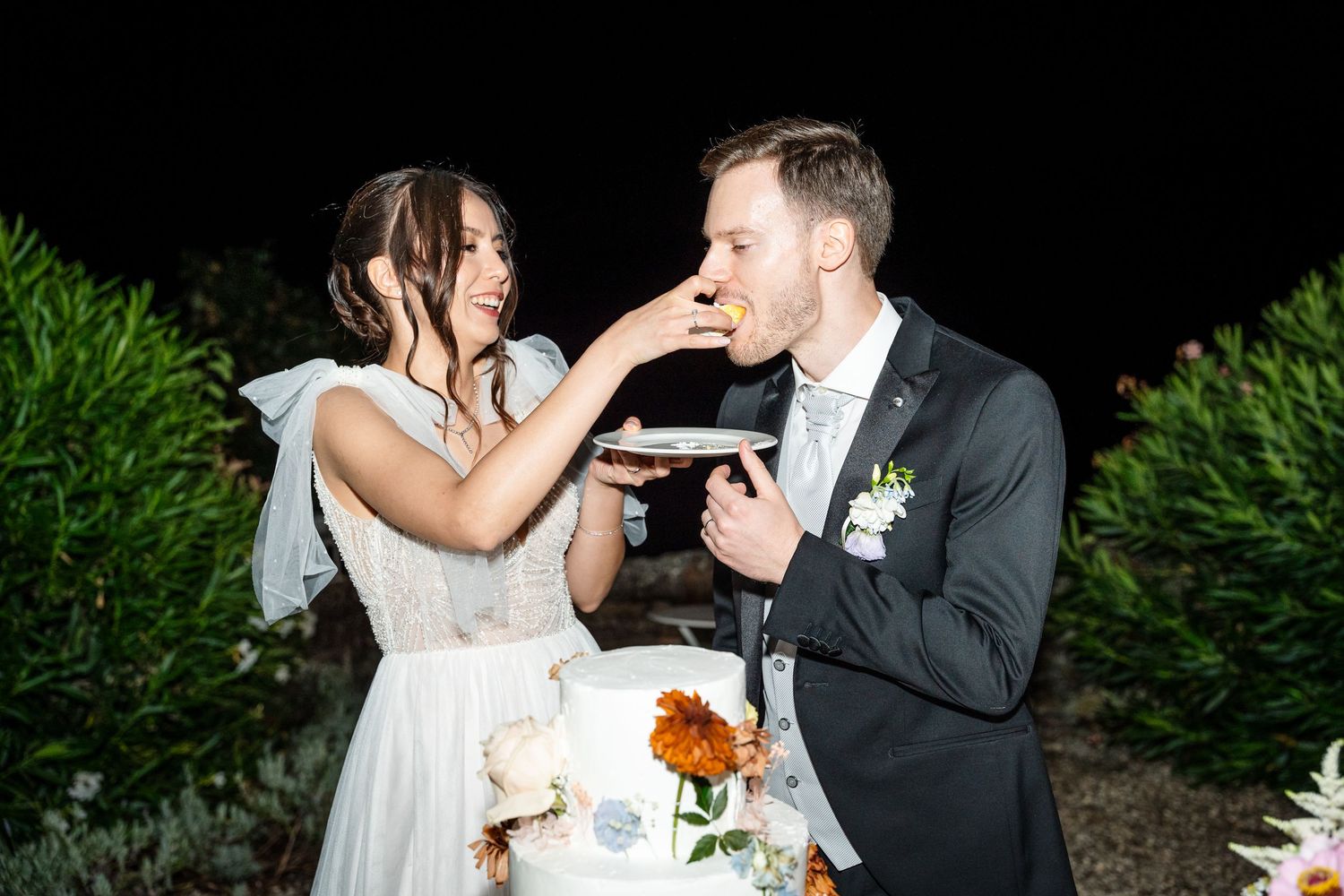 Bride feeding wedding cake to groom at outdoor night reception with white tiered cake.