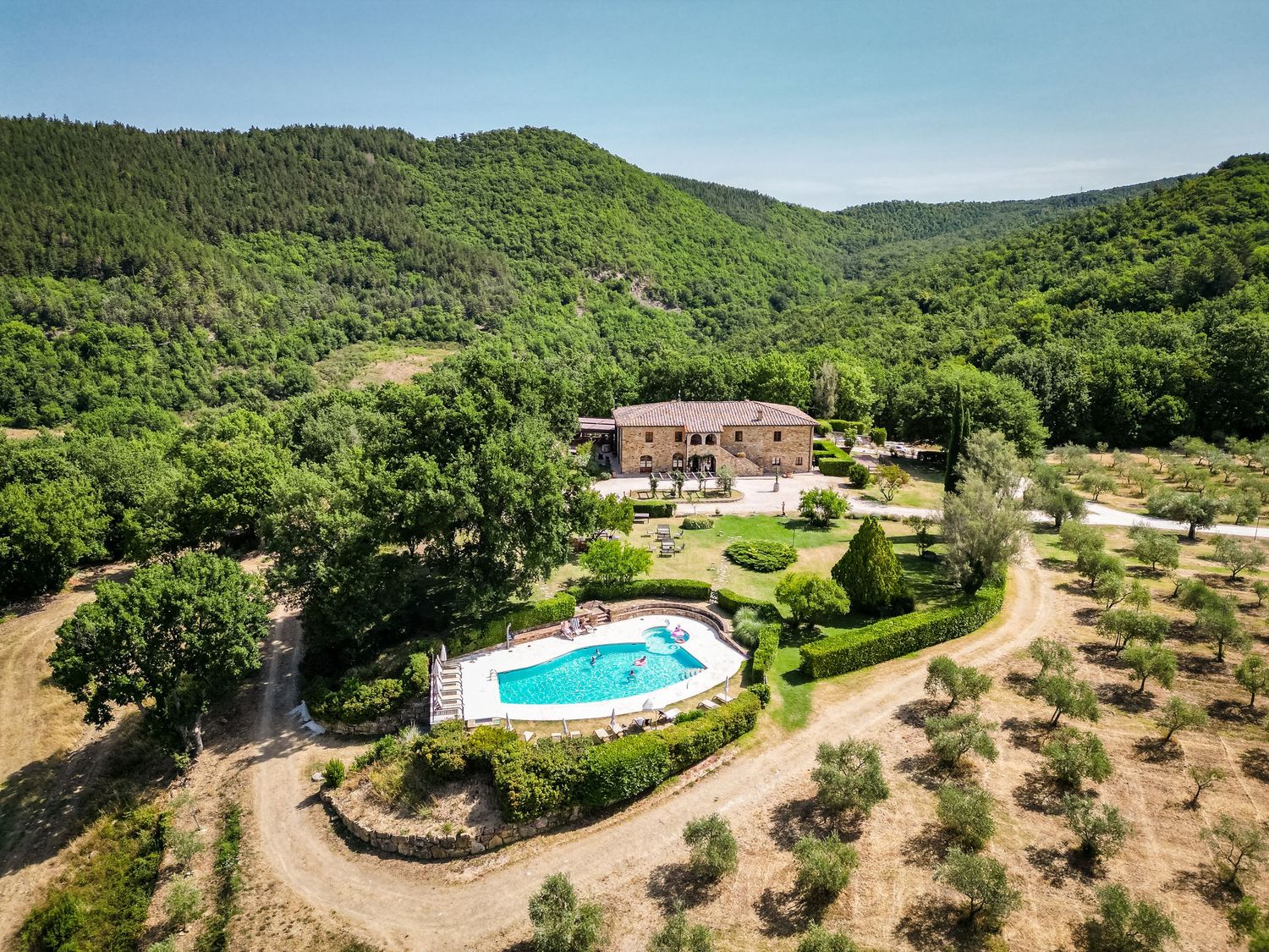 Aerial view of a Tuscan stone villa with pool surrounded by olive trees and lush green forested hills.