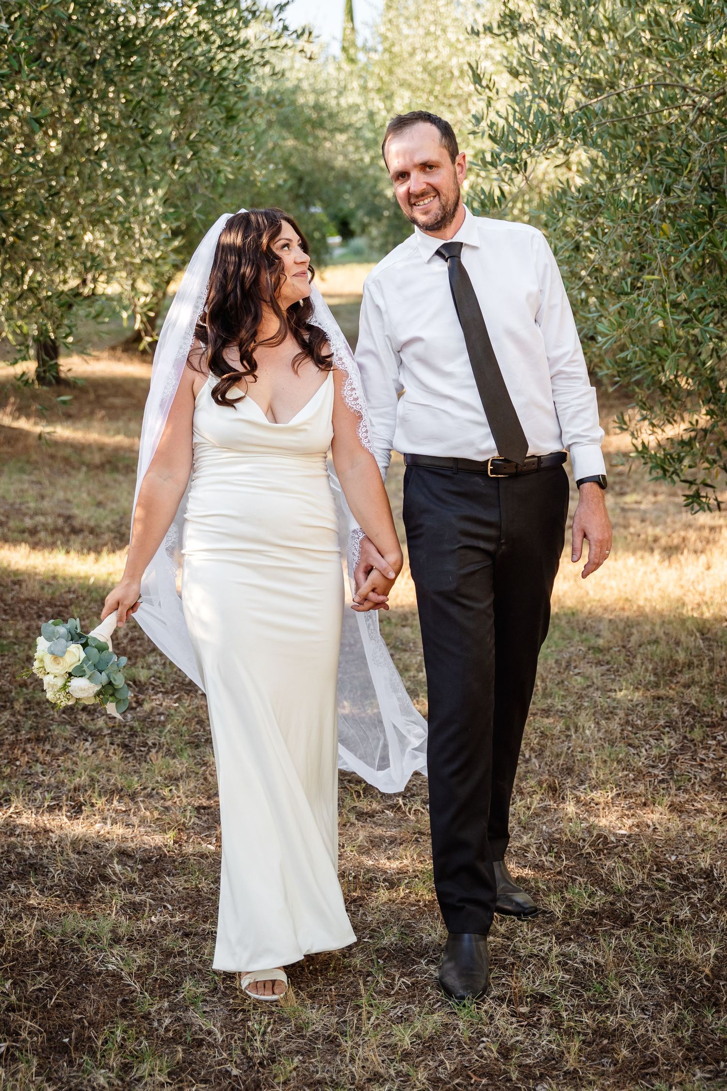 Newlyweds smile and walk hand-in-hand through a sun-dappled olive grove on their wedding day.
