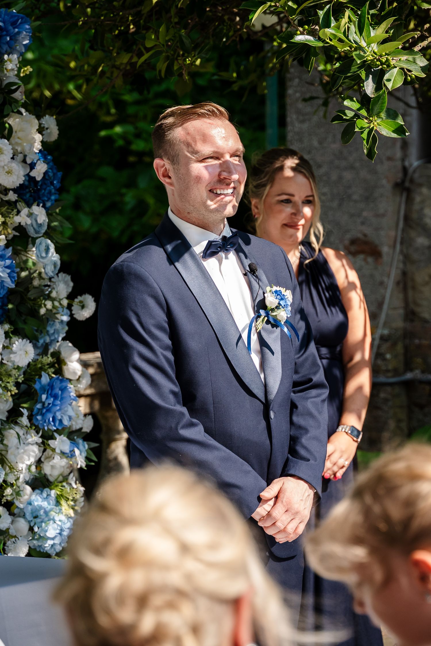 Groom in navy suit with boutonniere smiling at outdoor wedding ceremony with blue and white floral arch.