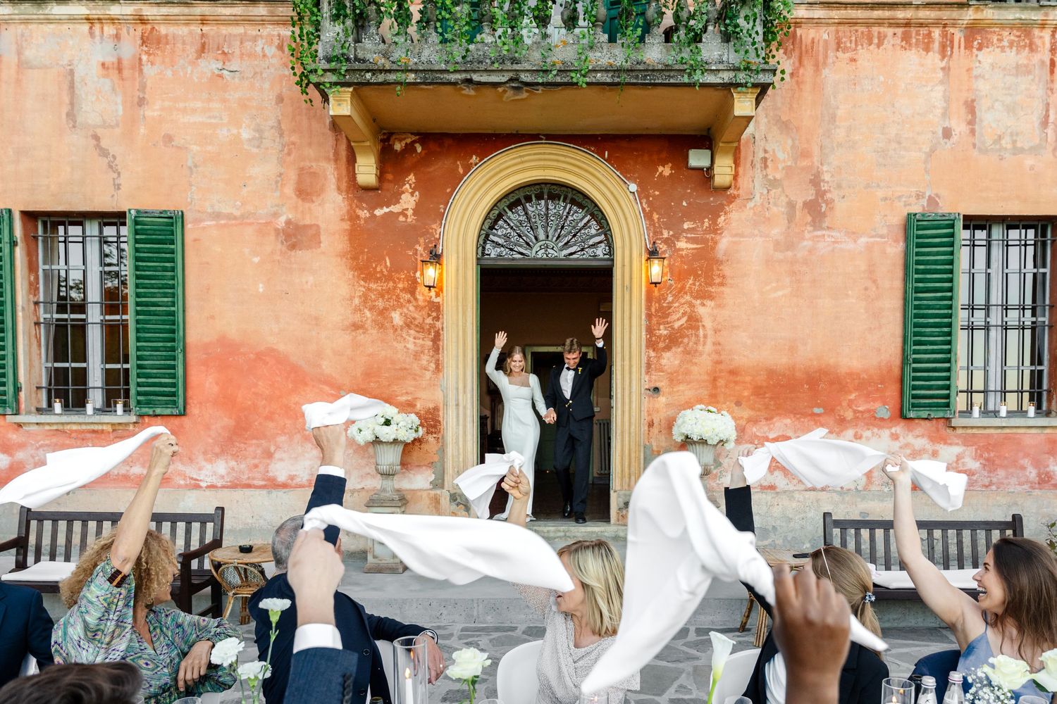 Newlyweds celebrate at a rustic Italian villa doorway as guests wave white ribbons in a joyful outdoor ceremony.