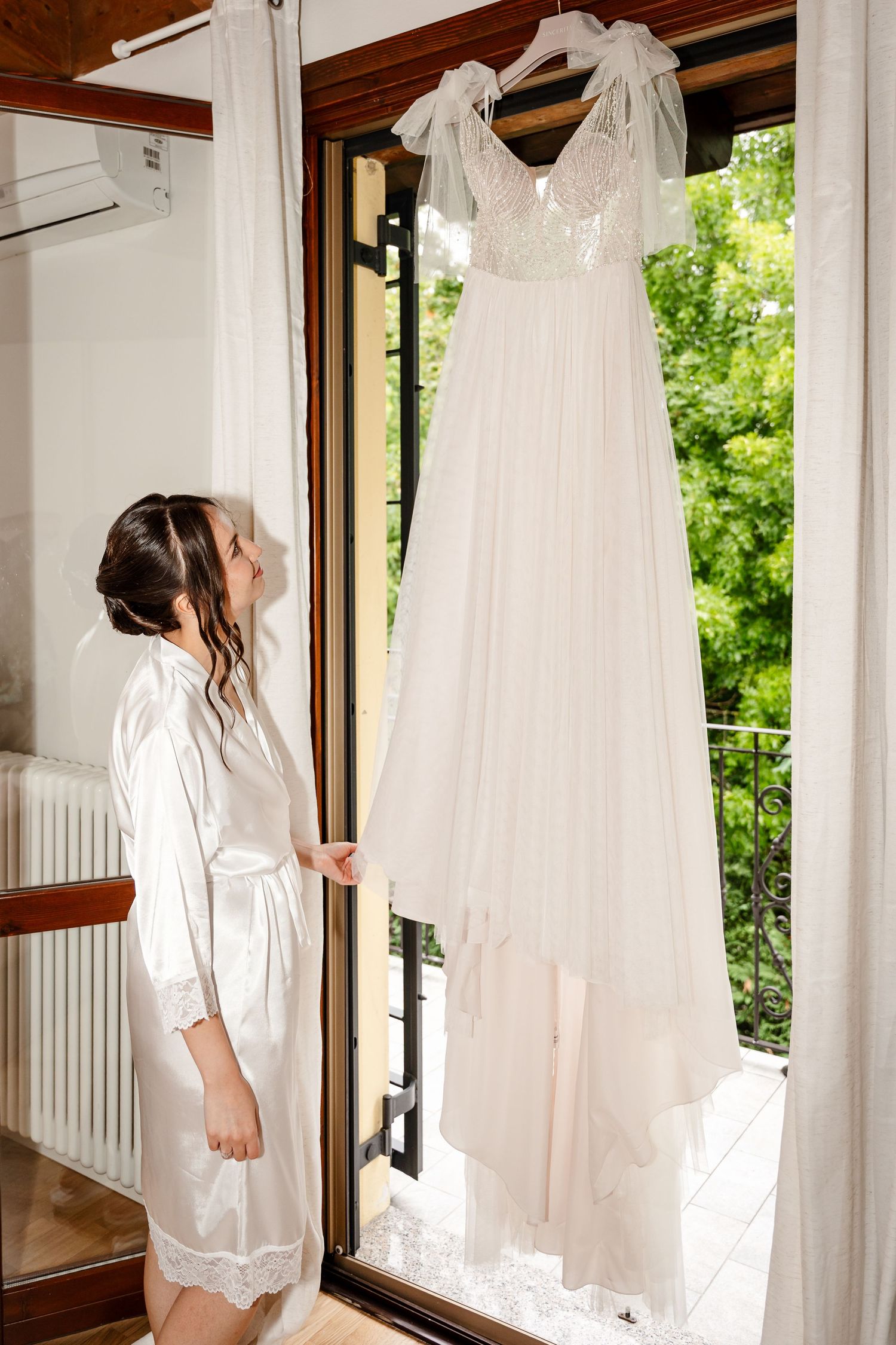 Bride in white satin robe admiring her wedding dress hanging in a sunlit doorway with garden views.