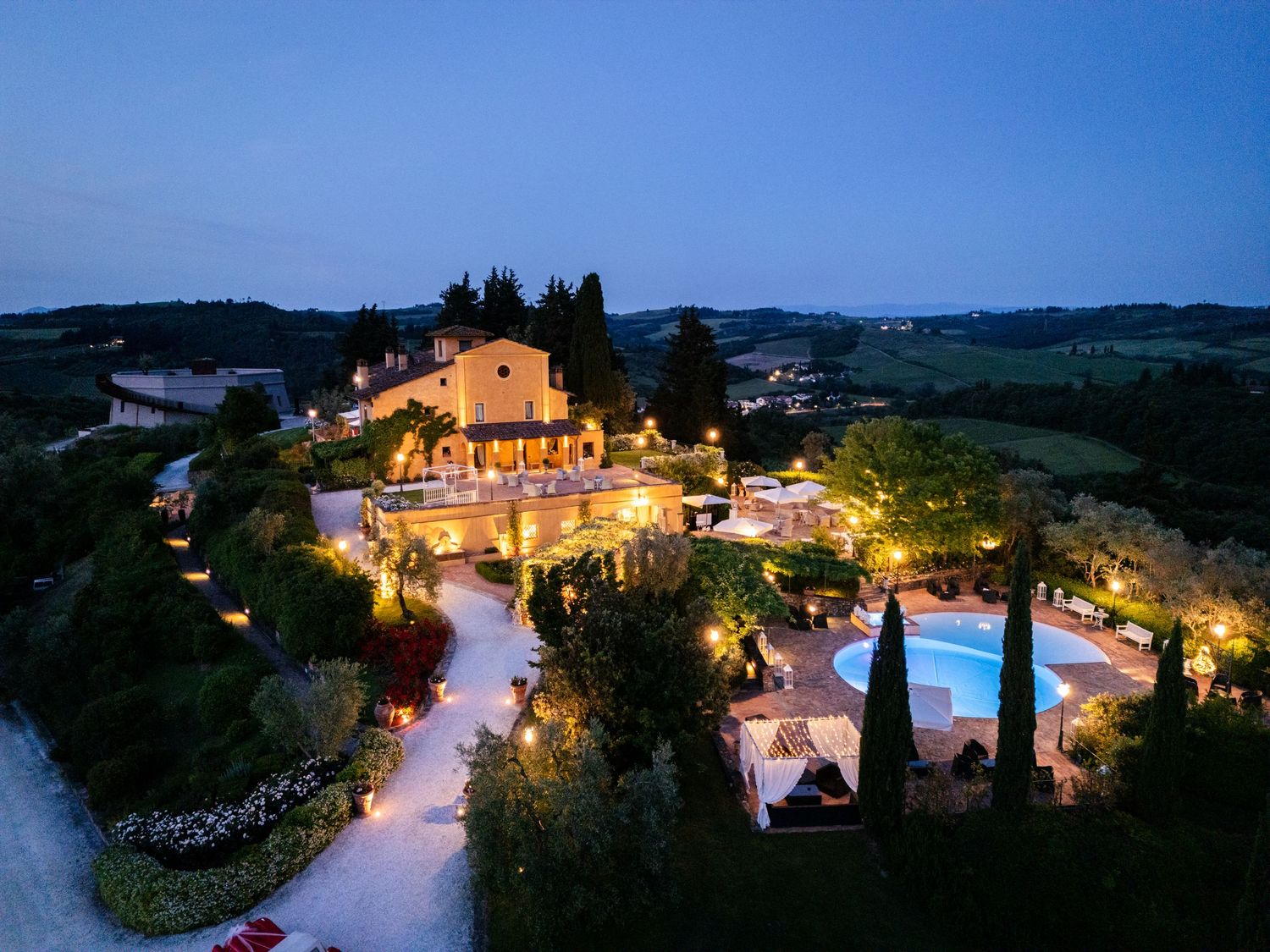 Aerial night view of a beautifully lit Tuscan villa with pool surrounded by rolling hills.