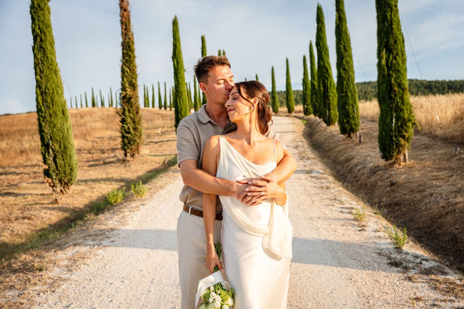 Couple embracing on a cypress-lined gravel path in Tuscany, bride in white gown holding bouquet, groom in tan suit.