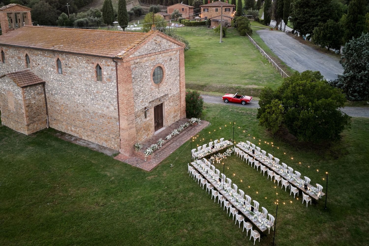 Aerial view of an elegant outdoor wedding reception setup beside a rustic Tuscan stone church with a red car on the road.