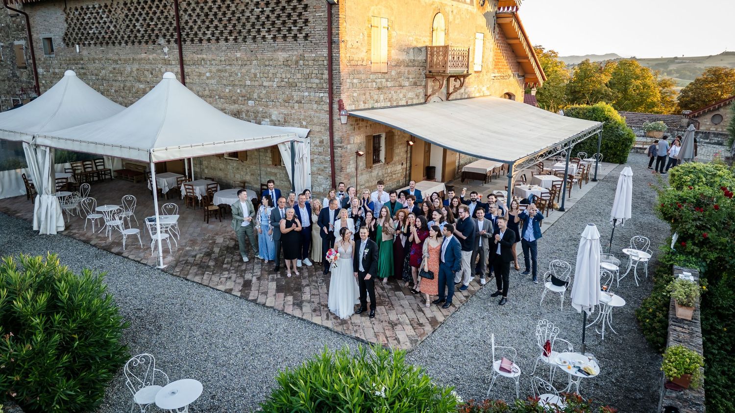 Large wedding group photo outdoors at elegant Italian venue with white canopy tents and vineyard views at golden hour.