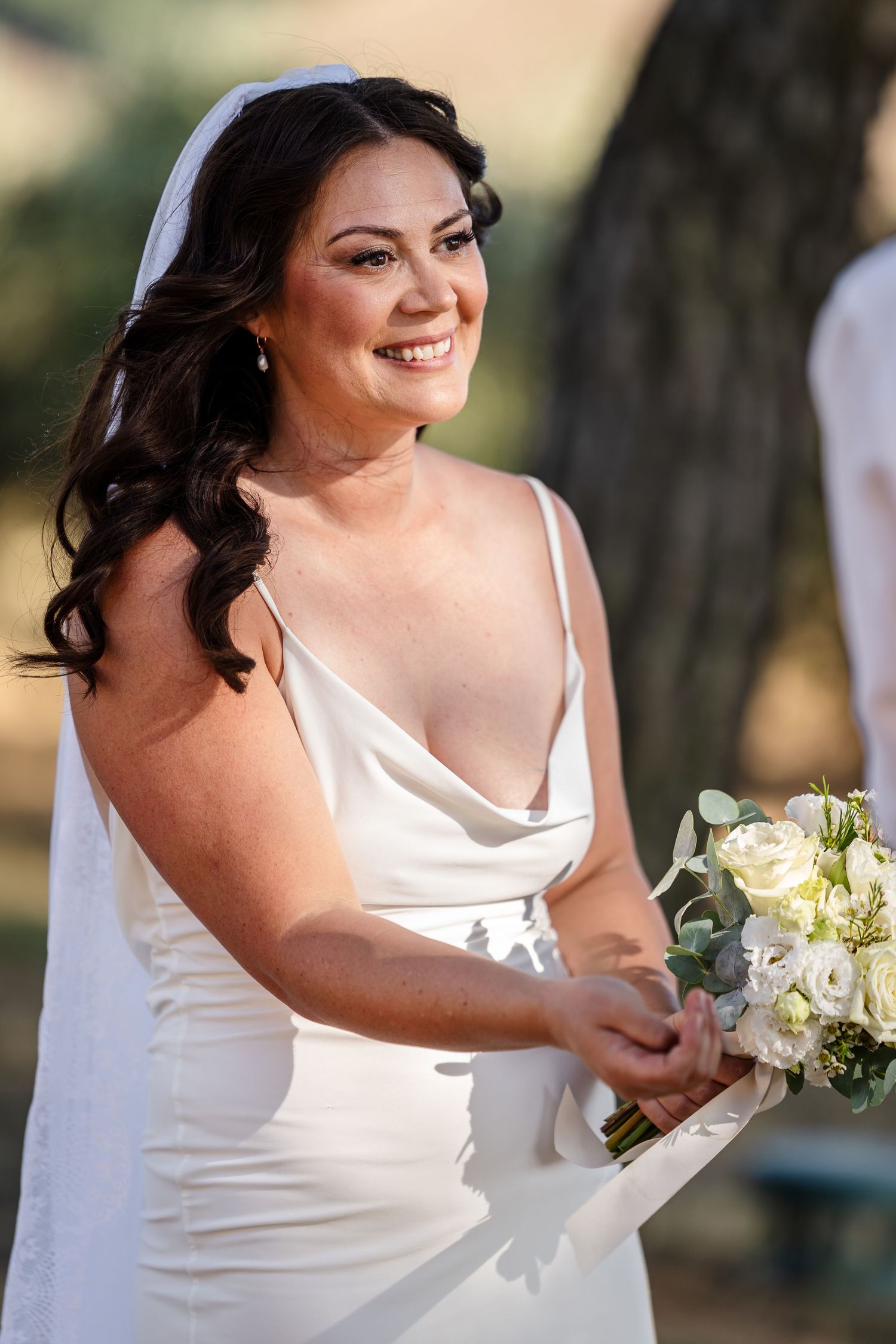 Smiling bride in white satin cowl-neck gown holding white floral bouquet outdoors.
