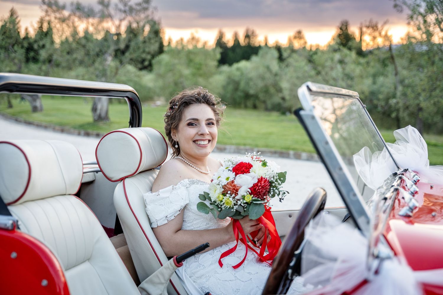 Smiling bride in white dress holding red roses bouquet seated in a vintage red convertible car.