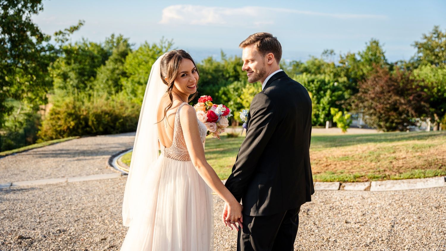 A couple in formal wedding attire embraces on a gravel path during their outdoor photoshoot.