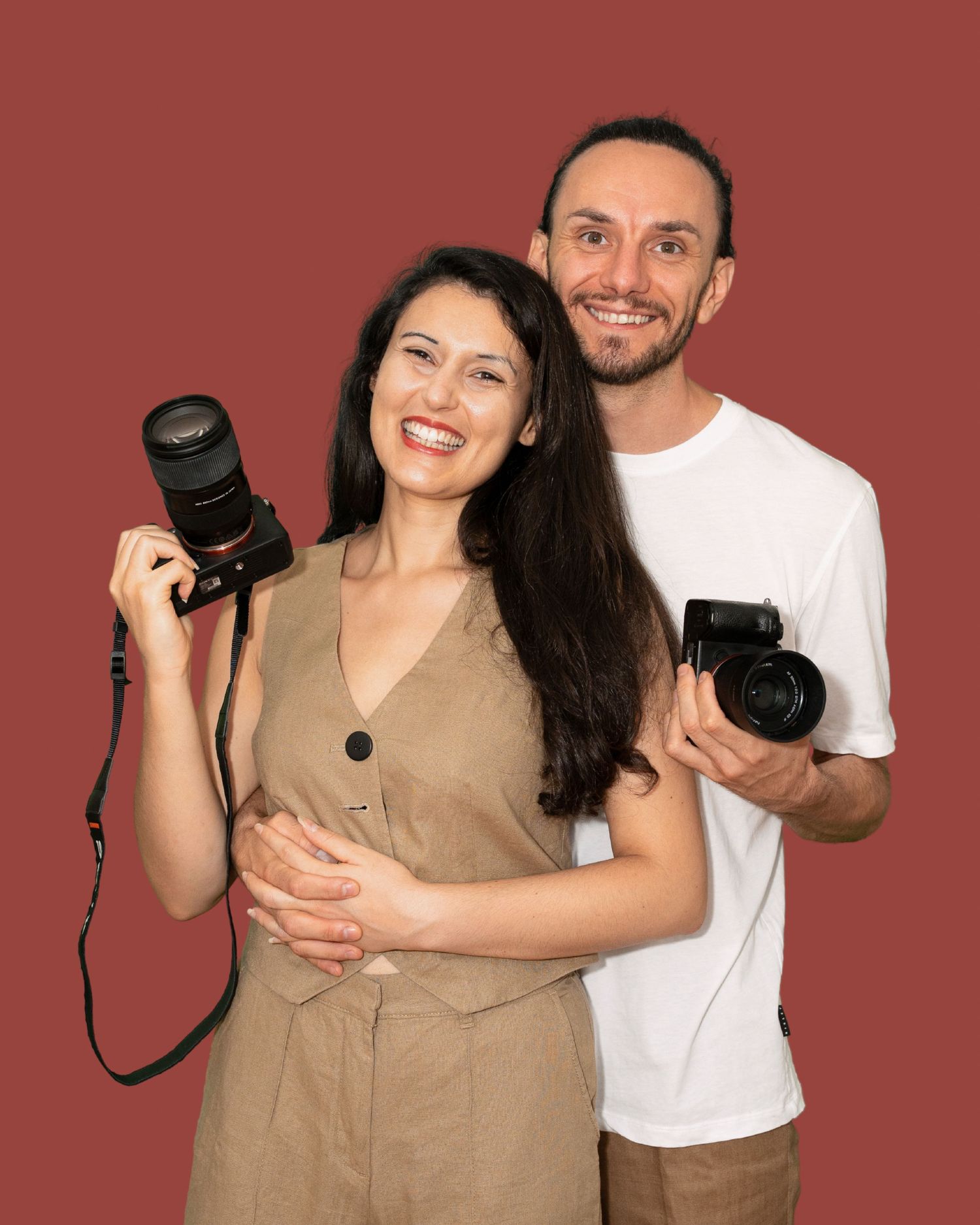Two photographers holding professional cameras pose together against a brown background.