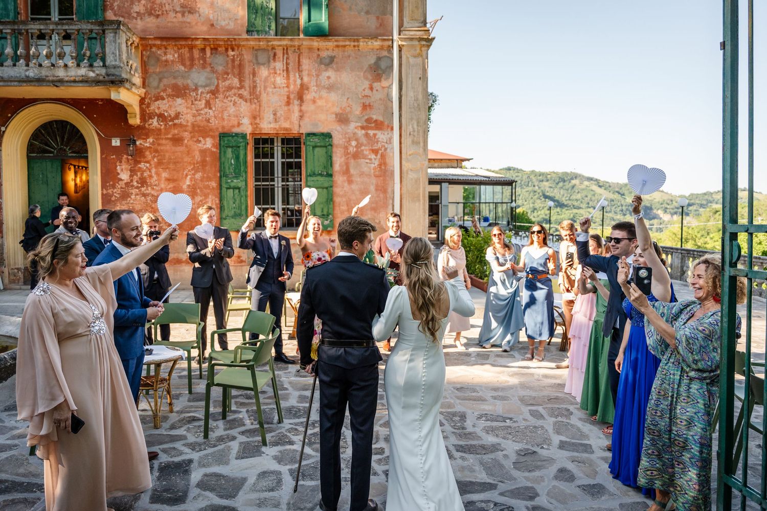 Bride and groom celebrated by wedding guests outside a rustic Italian villa on a sunny day.