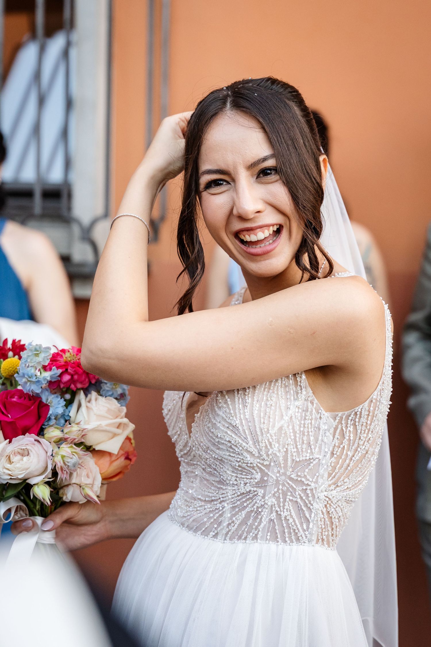 Joyful bride in beaded wedding gown laughing while holding colorful bouquet outdoors.