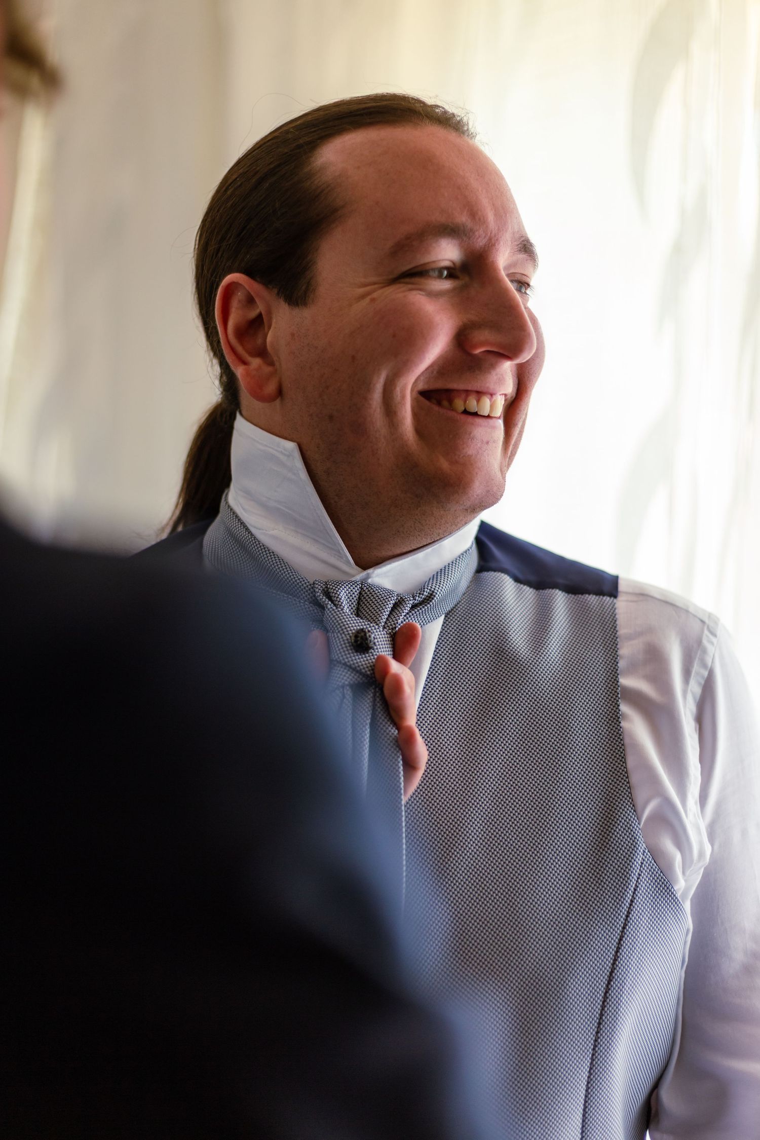 Smiling groom with long hair getting dressed in a blue suit and cravat tie before his wedding.