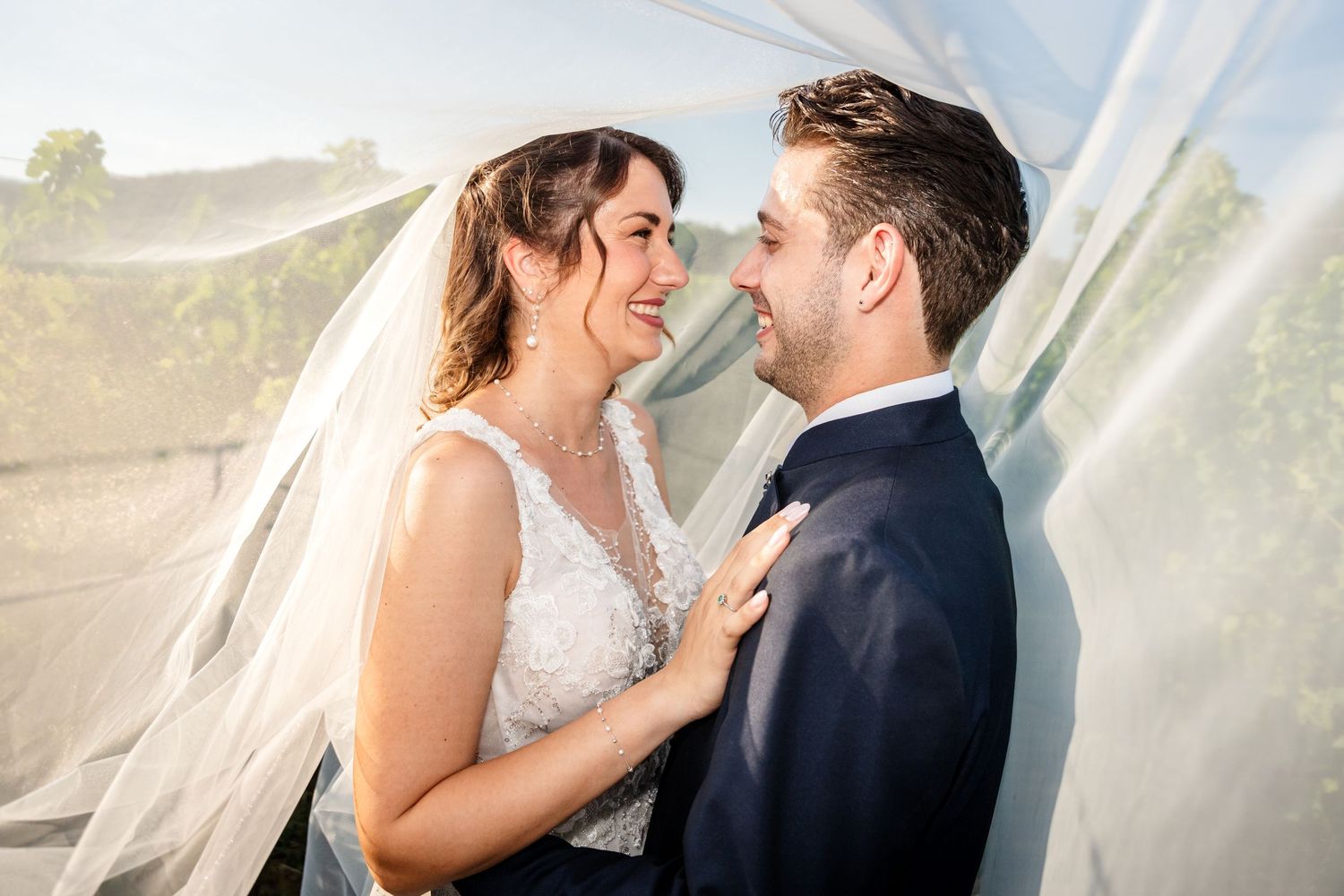 A bride and groom share a joyful moment under a flowing veil at their outdoor wedding ceremony.