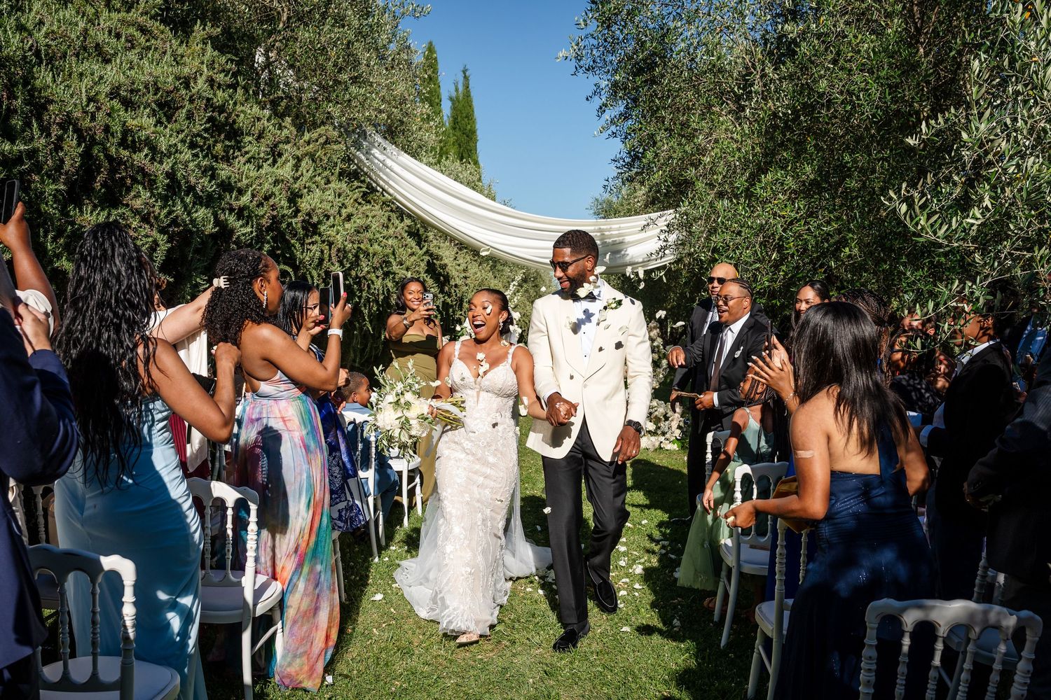 Joyful newlyweds walk down outdoor aisle surrounded by celebrating guests at a sunny garden wedding ceremony.