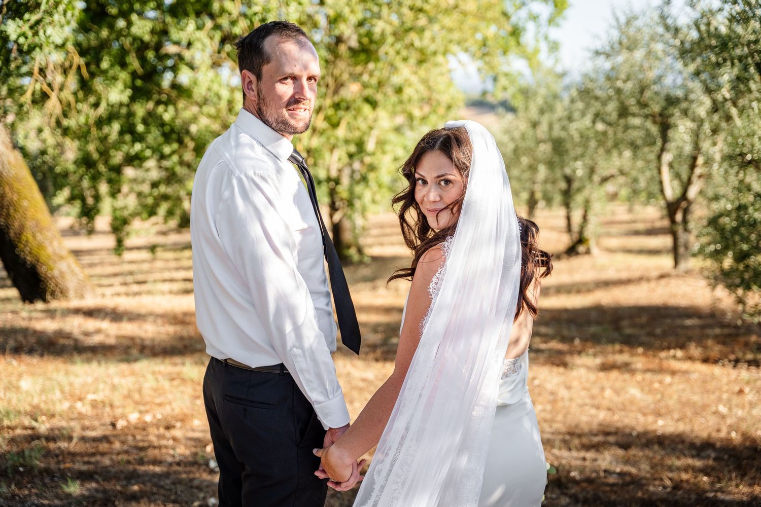 A bride and groom hold hands and smile outdoors among olive trees on their wedding day.