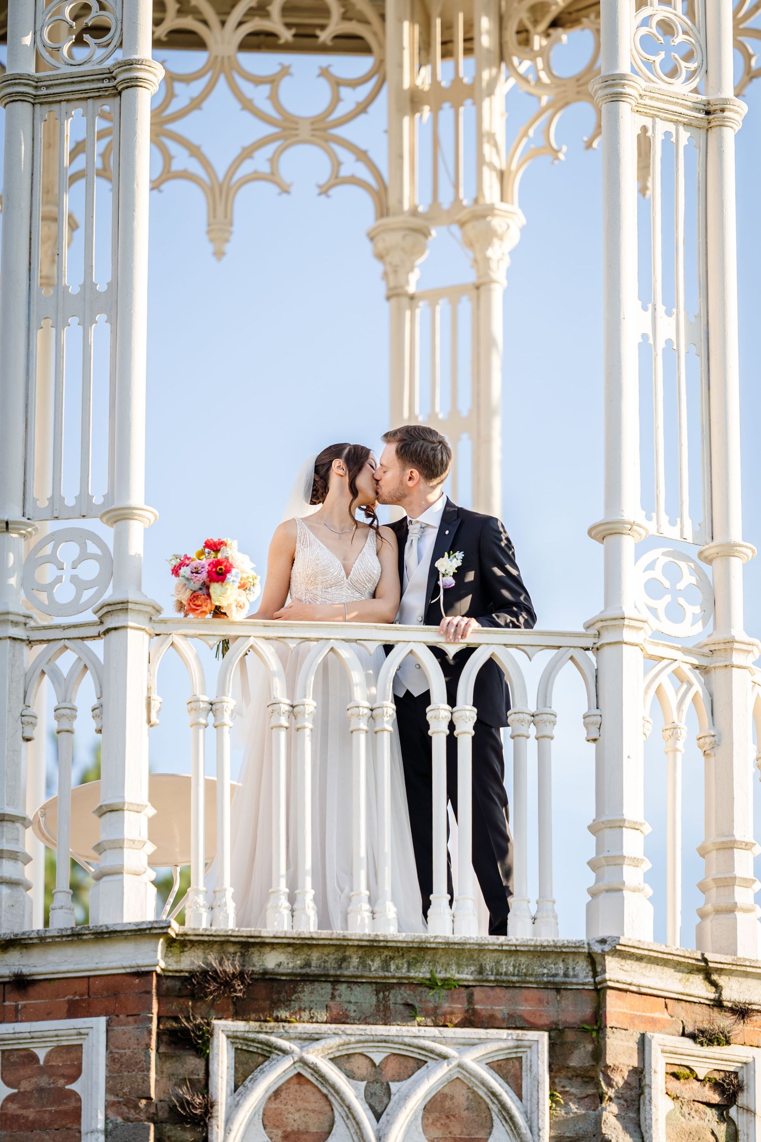 Bride and groom share a romantic kiss on an ornate white Gothic gazebo balcony, bride holding colorful bouquet.
