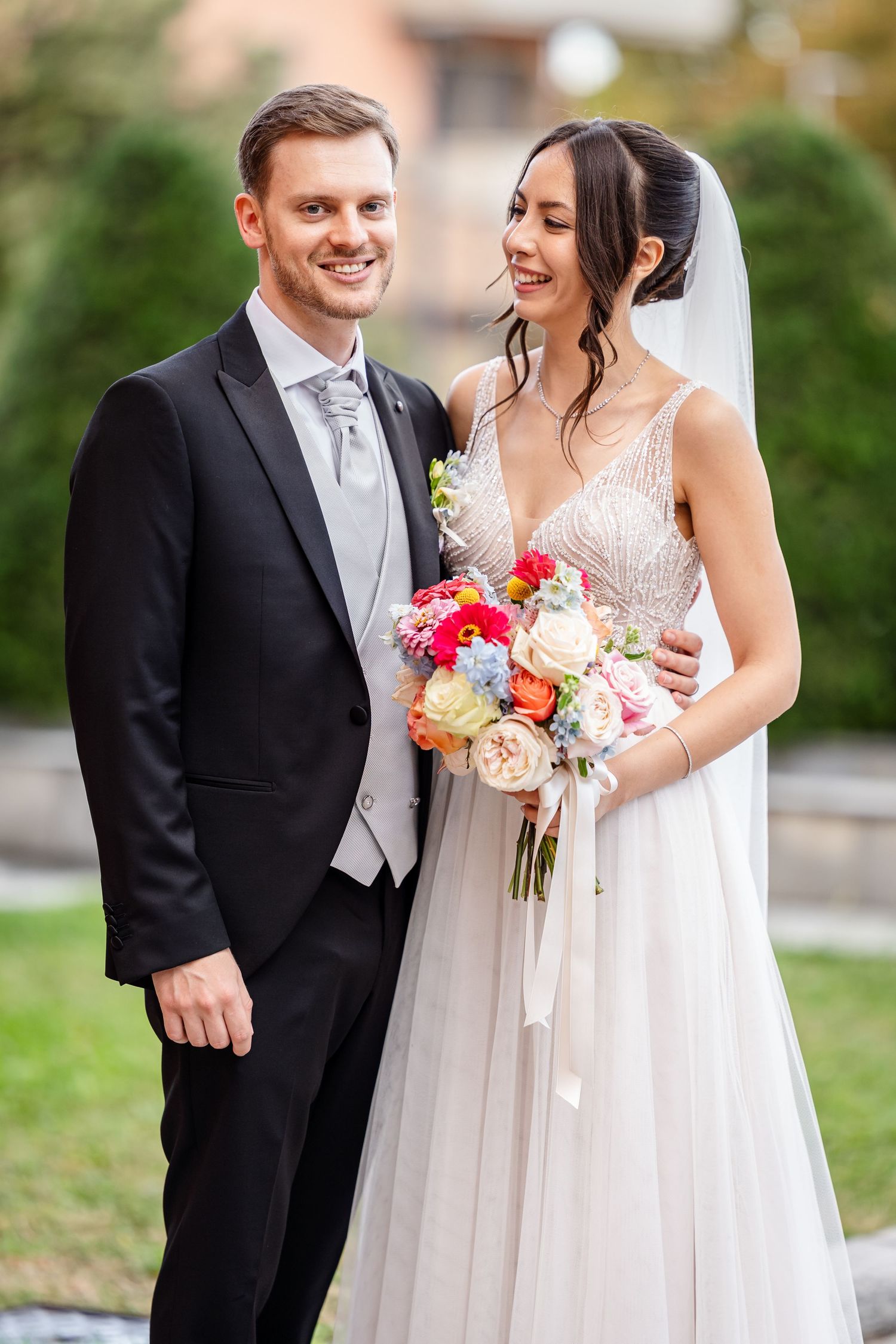 Smiling bride and groom pose together outdoors, bride holding colorful bouquet in elegant white gown and veil.