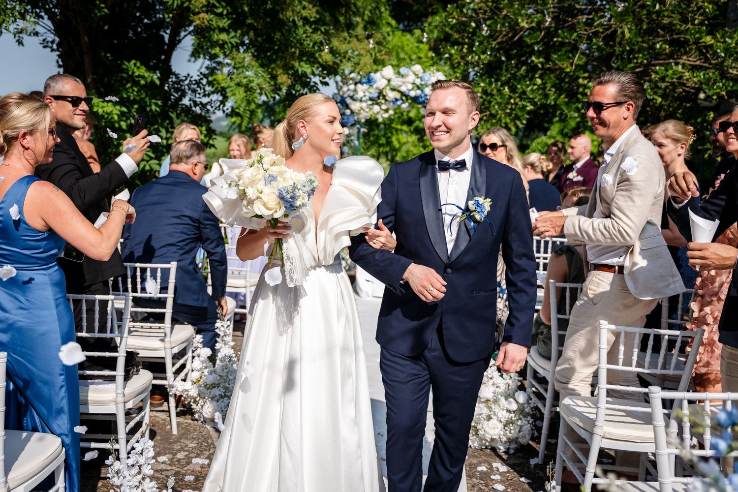 Bride and groom walk down outdoor aisle as guests celebrate with confetti at elegant wedding ceremony.