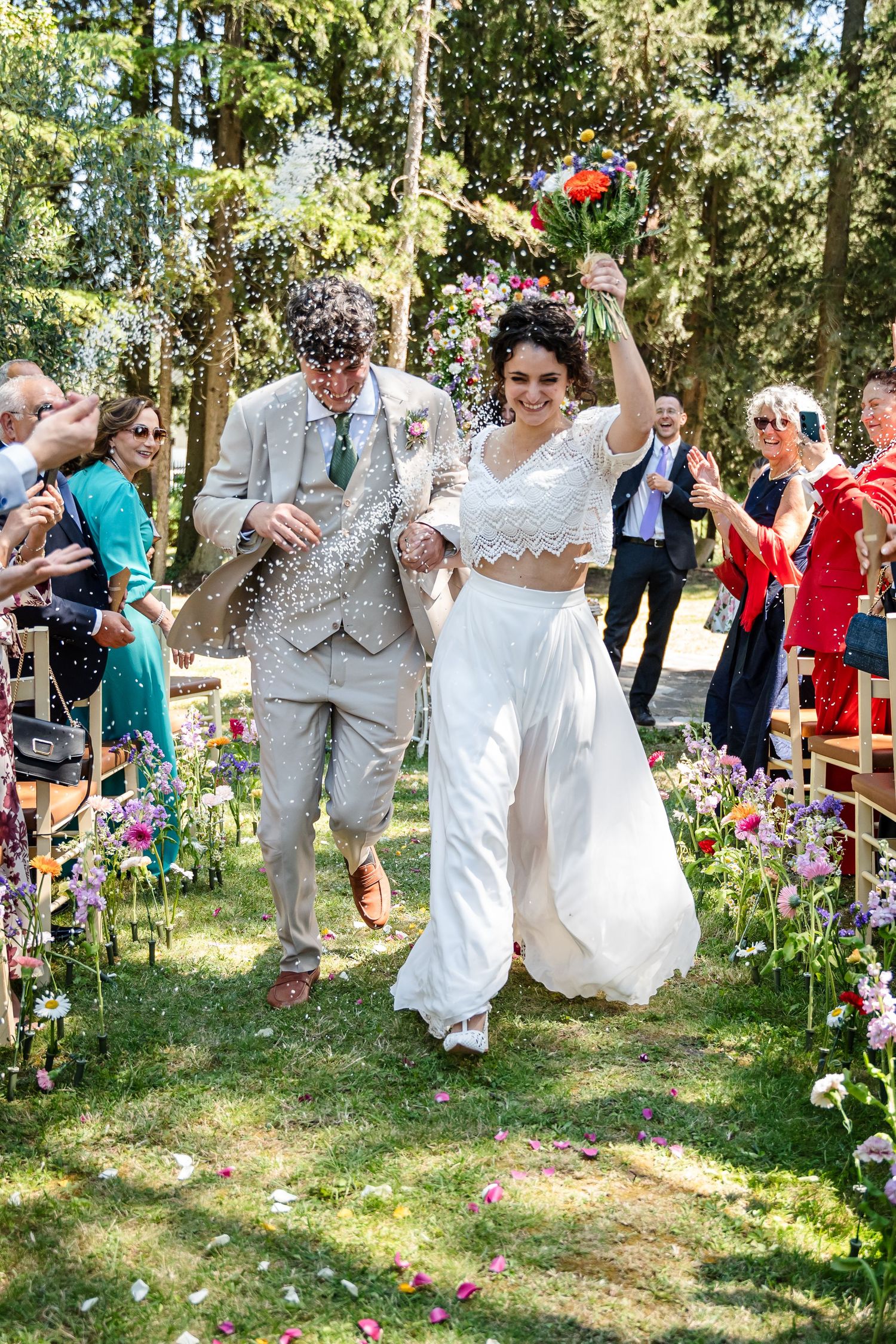 Joyful newlyweds walk down outdoor aisle as guests throw confetti at a colorful garden wedding ceremony.