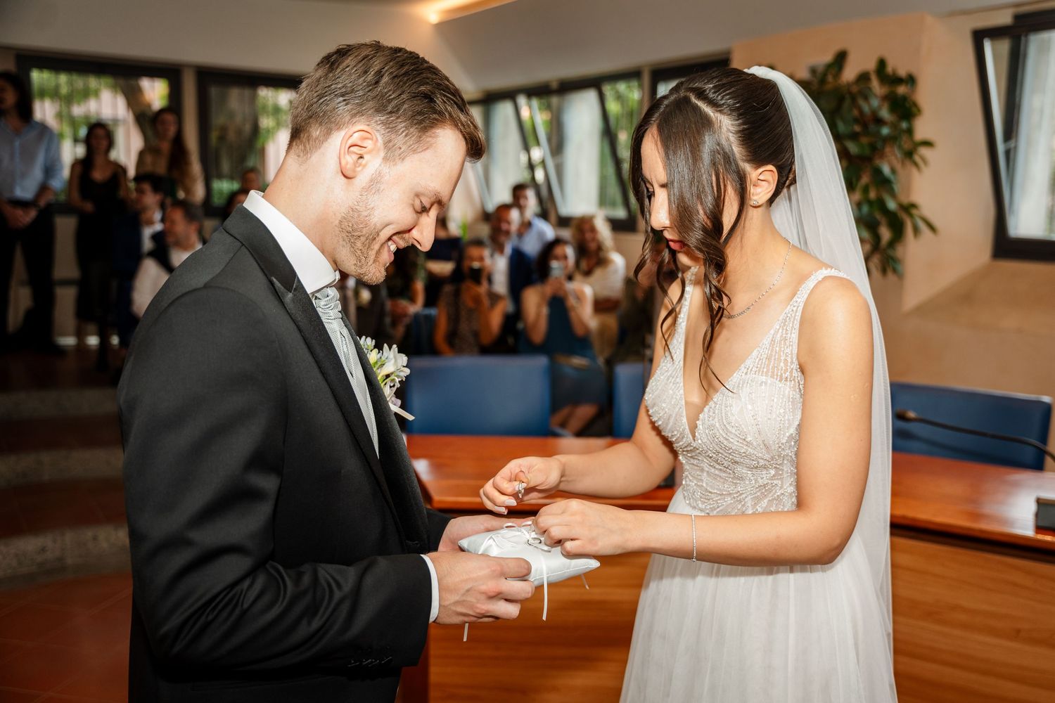 Bride placing wedding ring on groom's finger during indoor ceremony.
