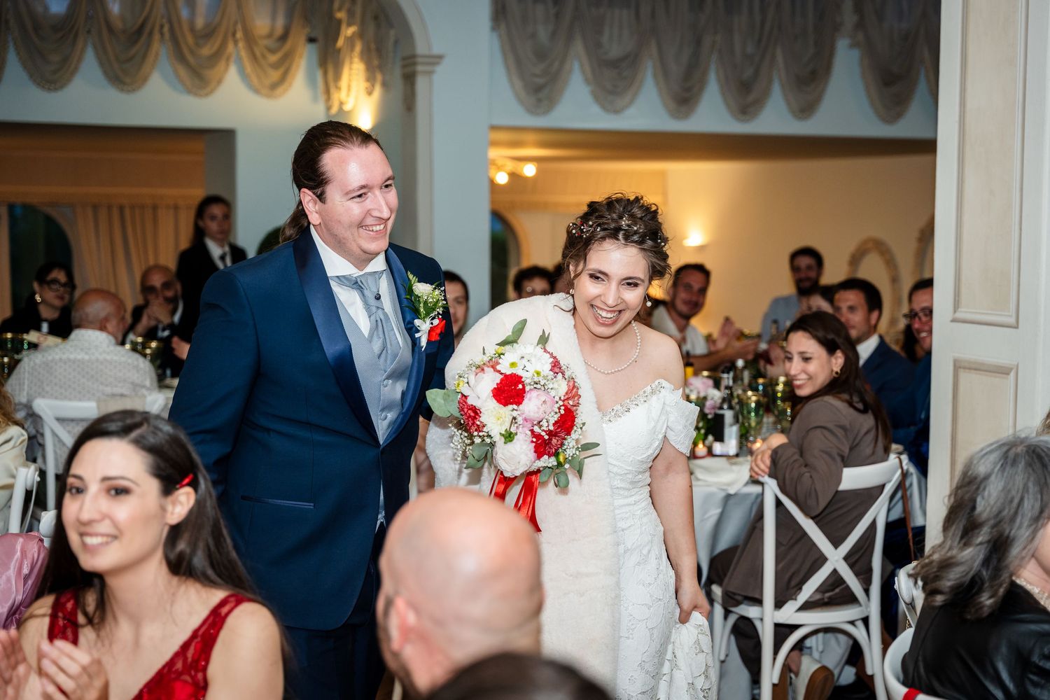 Bride and groom enter wedding reception hall smiling, bride holding red and white bouquet.