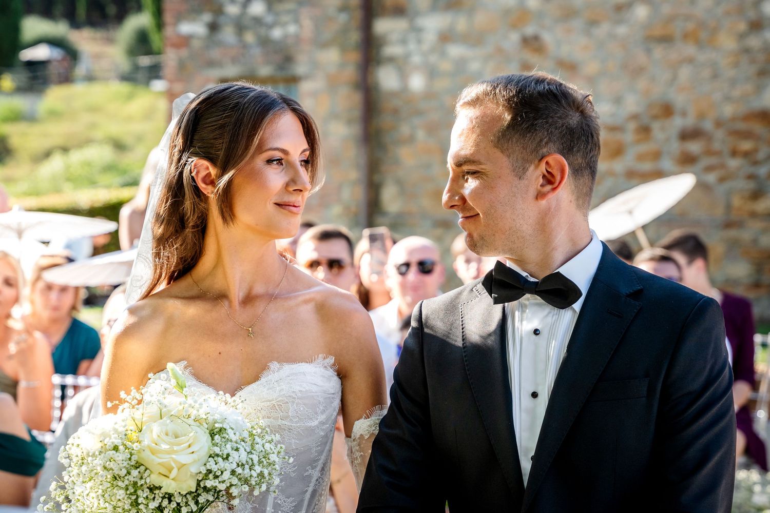 Bride and groom exchanging smiles at outdoor wedding ceremony, bride holding white bouquet, groom in black tuxedo.