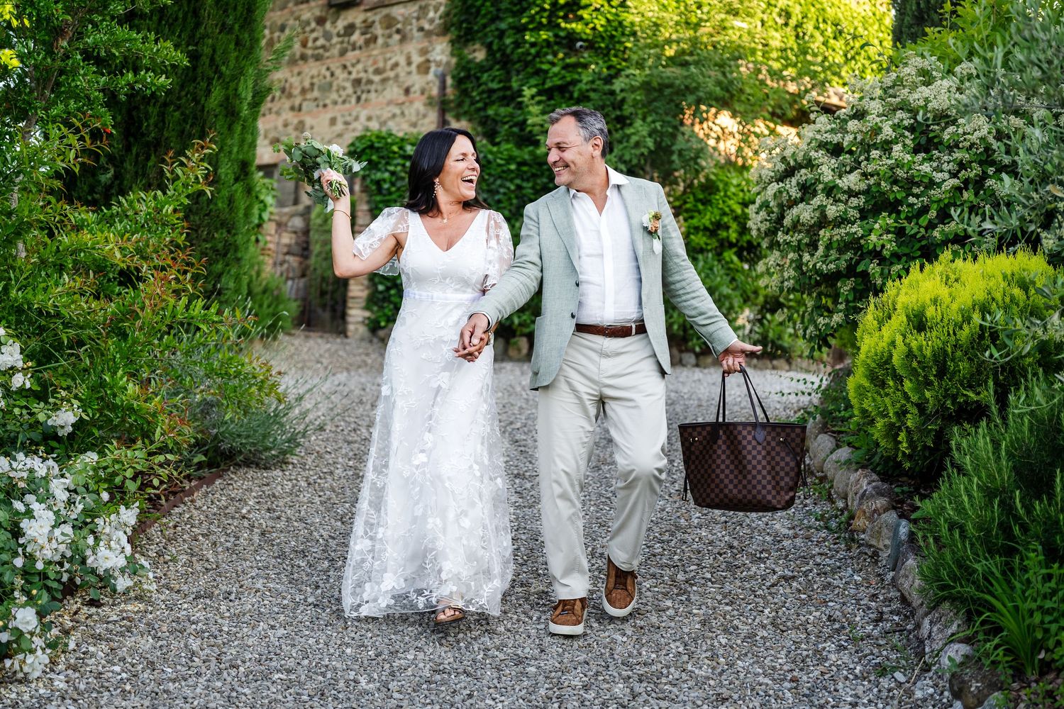 A couple in wedding attire walks along a garden path while holding hands and carrying a picnic basket.