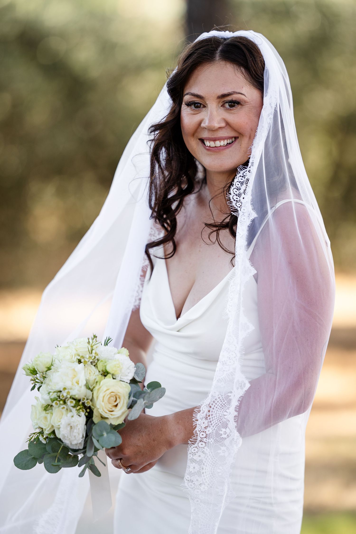 Beautiful bride in white cowl-neck gown and lace-trimmed veil poses with white rose bouquet outdoors.