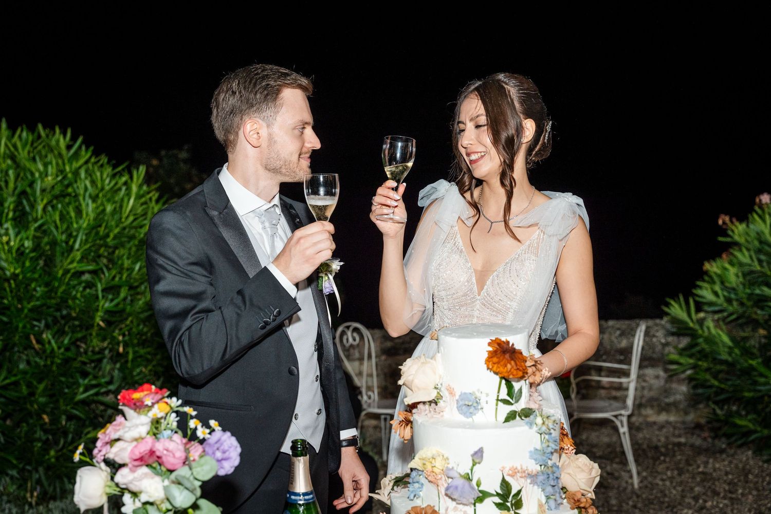 Bride and groom toasting with champagne glasses beside a floral wedding cake at night.